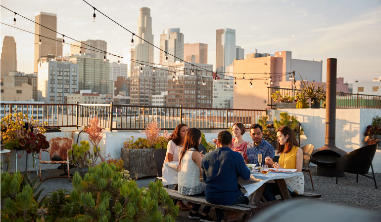 A group of friends share a meal on the rooftop of an L.A. apartment building with a view of the city skyline in the distance
