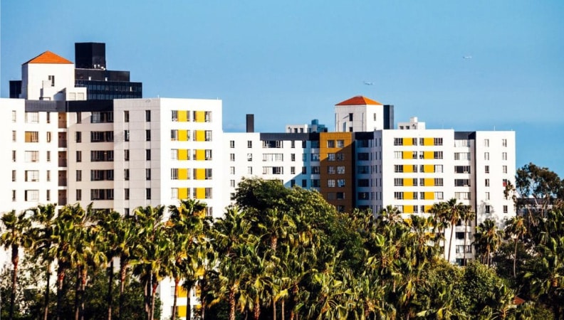 Large bunches of palm trees are growing in front of an apartment complex in the Los Angeles area. The apartment buildings are of varying heights and are painted white with yellow and brick accents.