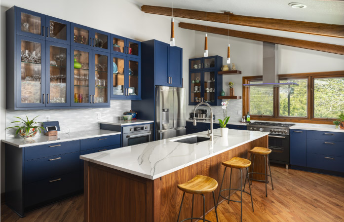 A newly remodeled kitchen with a marble-topped wooden island with a built-in sink, windowed cabinets painted in dark blue, and new chrome appliances. There are also modern pendant lights hanging from the ceiling above the island.
