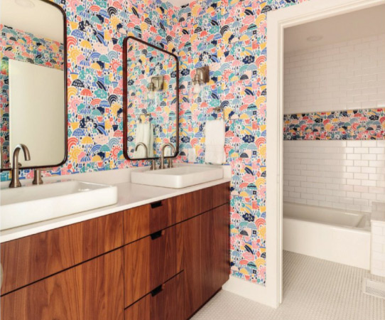 A newly remodeled bathroom with mid-century modern mirrors and vanity. The walls in the vanity room have been decorated with colorful wallpaper. The walls in the room with the bath are covered in white subway tiles with a single strip of matching wallpaper running horizontally in the middle. 