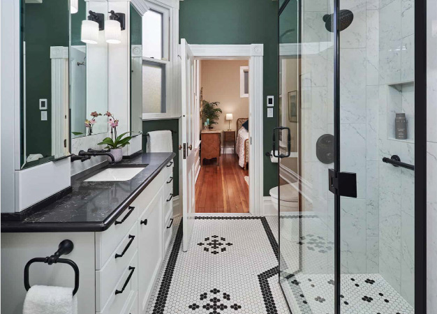 A newly remodeled bathroom with custom tilework on the floor and in the shower, a white vanity with a black marble top, and dark green painted walls with white trim.