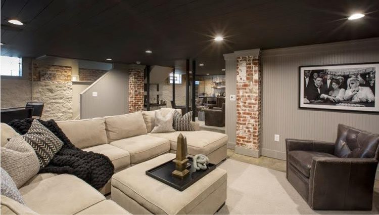 A remodeled basement featuring exposed brick and wood-paneled walls, black painted ceilings, recessed overhead lighting, and cozy furniture. The room’s decor includes a few statuettes on the ottoman and a photograph of old Hollywood actors on the wall.