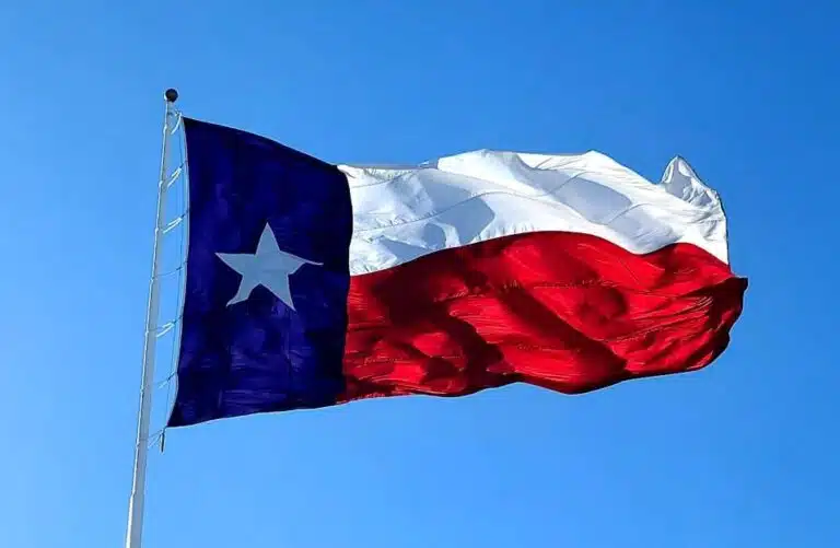 View of the Texas State flag blowing in the wind at the top of a flagpole in front of a clear blue sky.