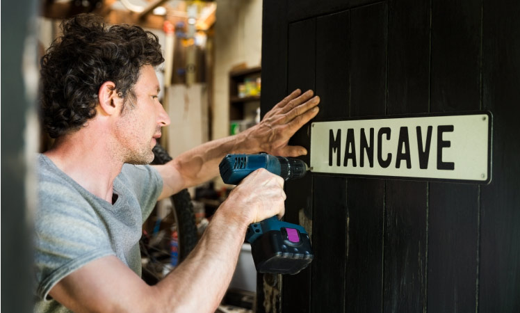 A man uses an electric drill to hang up a sign that says “MANCAVE” on the door of his garage, which he recently remodeled into a living space.