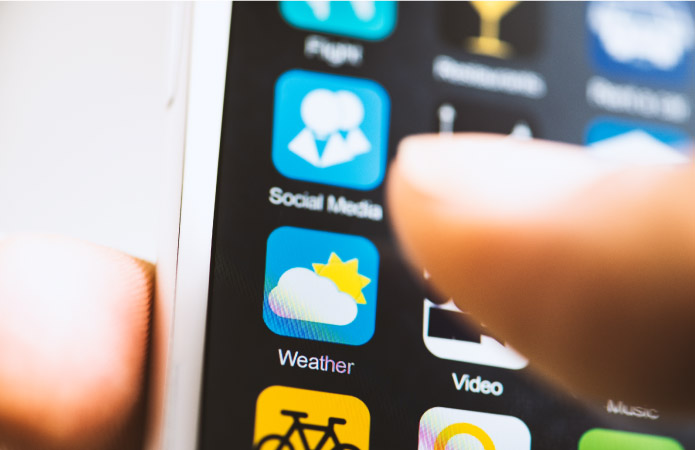 A close-up of a hand holding a smartphone. One finger is about to tap on the “Weather” app icon.