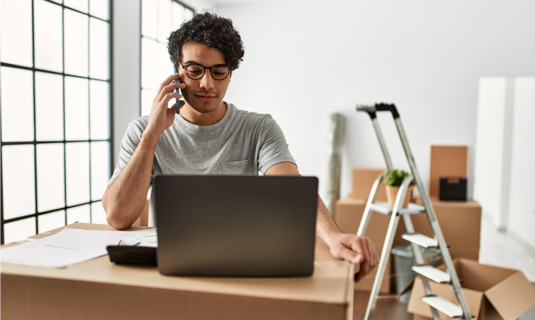 A man is sitting at a make-shift desk (i.e., a cardboard box) with his laptop and some papers. He’s on the phone setting up the utilities at his new place. There is a step-ladder and several other moving boxes behind him.