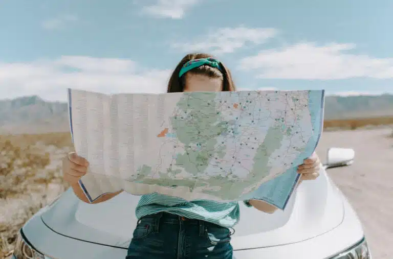 In the process of moving out of California, a young woman is parked on the side of an interstate, leaning on the hood of her car while looking over a map.