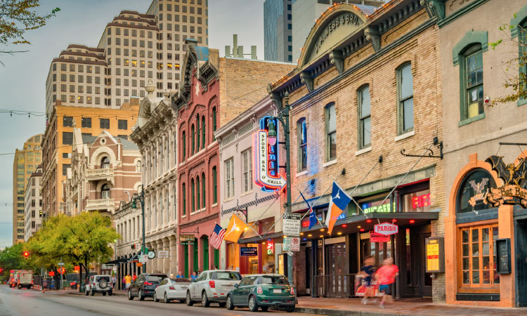 The entertainment district in Austin, Texas, during the day. People are walking along the sidewalk and cars are parked in the street. There are various flags hanging from a storefront, including the Texas state flag, the U.S. flag, and an orange Texas Longhorns flag.