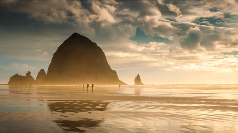 Several people are enjoying a stroll on Cannon Beach along the Oregon Coast at sunset. The sand is wet and reflective, and Haystack Rock appears to rise up in the distance. 