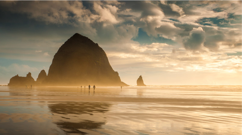 Several people are enjoying a stroll on Cannon Beach along the Oregon Coast at sunset. The sand is wet and reflective, and Haystack Rock appears to rise up in the distance.