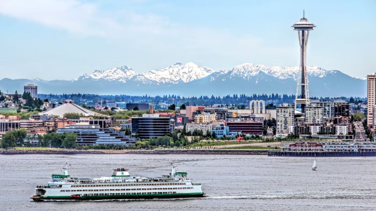 Daytime view of Seattle, Washington, featuring a ferry crossing Puget Sound, the Space Needle, and the Cascade Mountains in the distance.