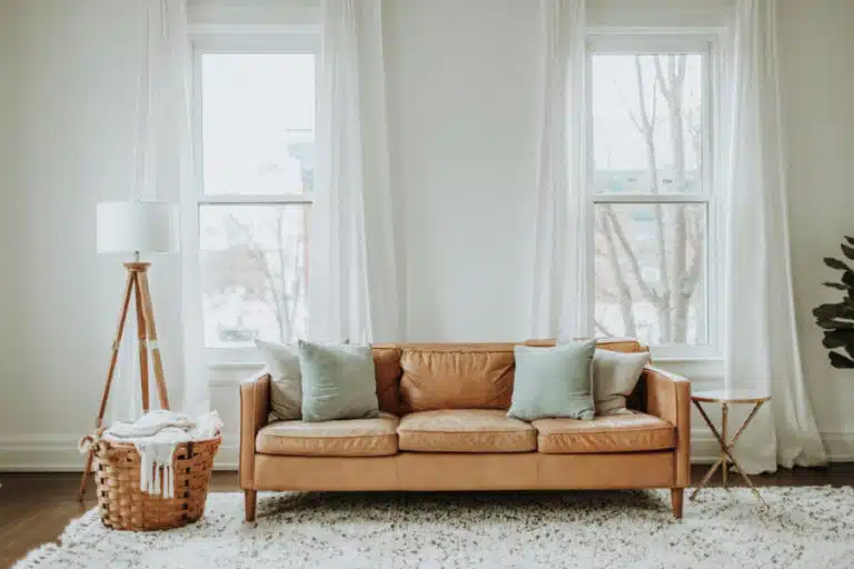 A simple minimalist living room featuring white walls, curtains, and an area rug, as well as a tan leather couch with a complimentary wooden lamp and side table. 