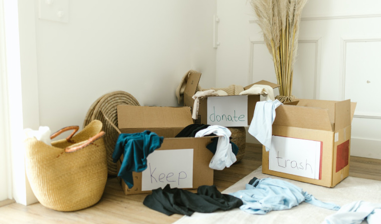 Three cardboard boxes are in the corner of a room. They’re labeled with paper signs that read “keep, ”donate,” and “trash,” respectively. There are a few clothing items on the floor and some that are hanging off the sides of the boxes. 