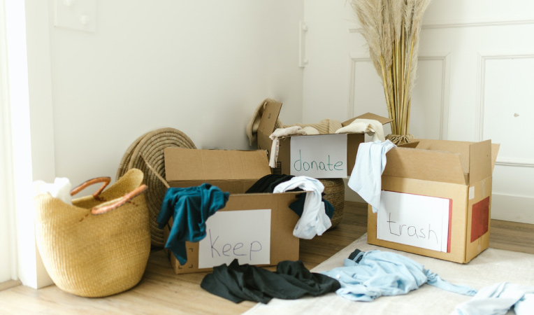 Three cardboard boxes are in the corner of a room. They’re labeled with paper signs that read “keep, ”donate,” and “trash,” respectively. There are a few clothing items on the floor and some that are hanging off the sides of the boxes.