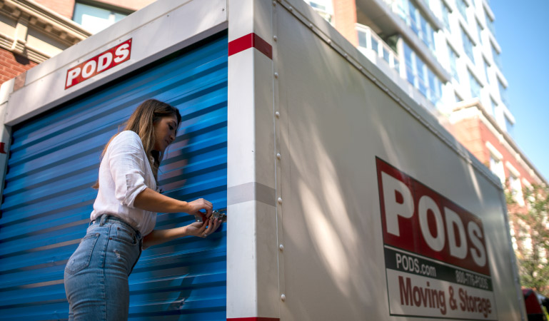 A woman is locking the door on her PODS portable storage container. The contains is positioned on the side of a street in a big city. She’s using it to store items that are cluttering her home but that she doesn’t want to get rid of.