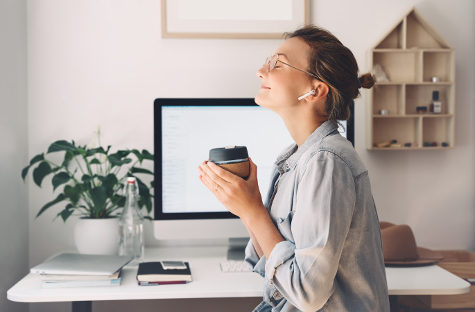 A woman is listening to music through earbuds while she enjoys a warm drink in her minimalist office. Her eyes are closed and she’s smiling.