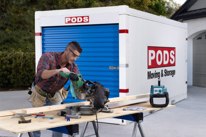 A man is using a saw to cut a wooden board in his driveway. Behind him is a PODS portable storage container, which he rented to use as temporary on-site storage during a home remodeling project.