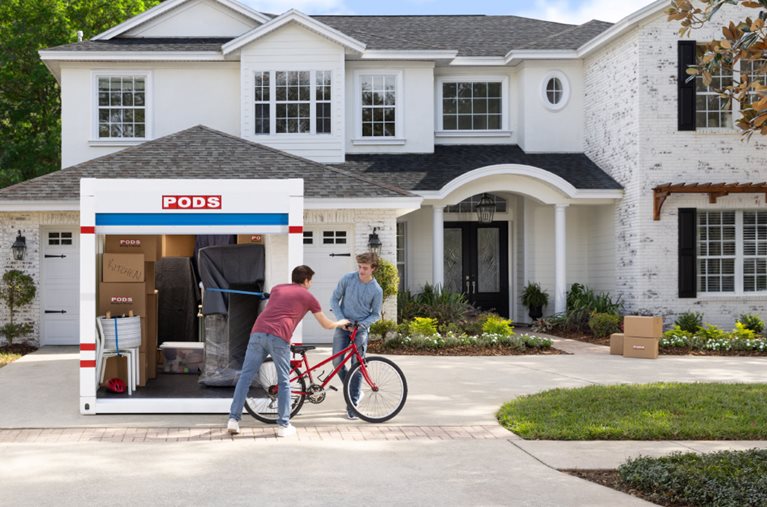 After renting a container for moving, a father is handing his son a red bicycle as they unload the container in the driveway of their new house.