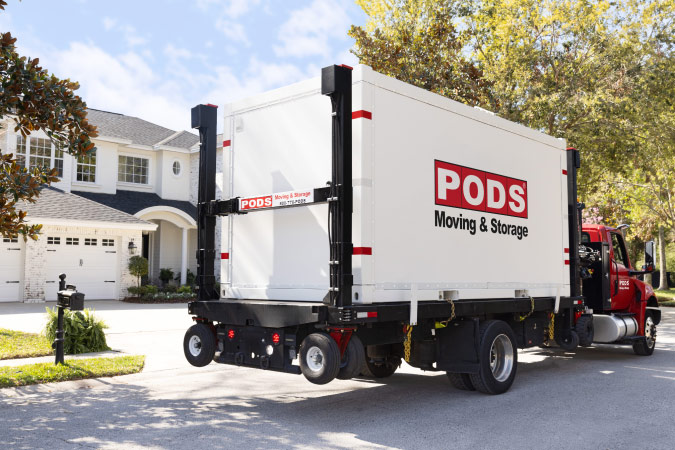A PODS truck with a PODS portable moving container on its bed is parked in front of a residential home.