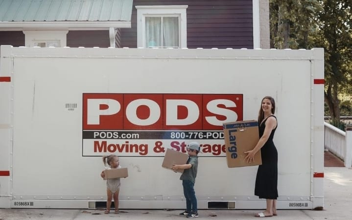 Katherine Tuttle and her two young children are standing in front of their PODS container, each holding a moving box.