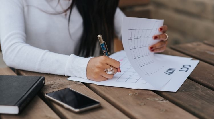 A woman is sitting at a wooden table with a print-out calendar. She’s marking the dates of her upcoming move to help keep track of when her portable moving container will be delivered and other details of her move.