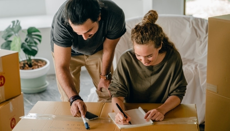 A couple is carefully labeling moving boxes and making a list of the items they're packing to track their belongings during their move.