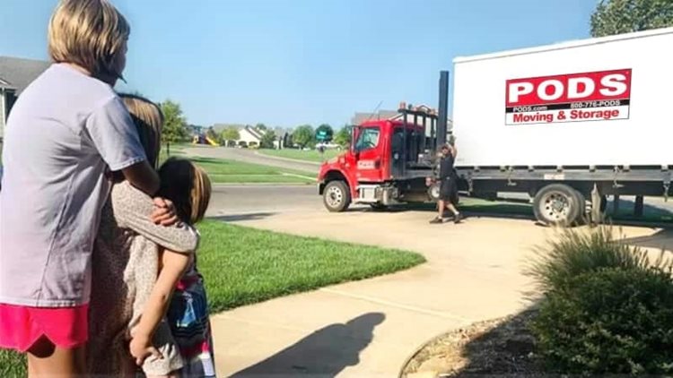 A woman and her two young children stand in their front walkway, watching the PODS truck driver prepare to transport their PODS portable moving and storage container to their new home.