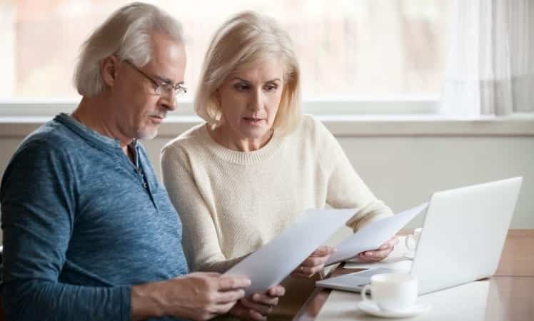A senior couple is sitting at a table with their laptop and some loose papers, reviewing moving insurance policies to find the right one for them.