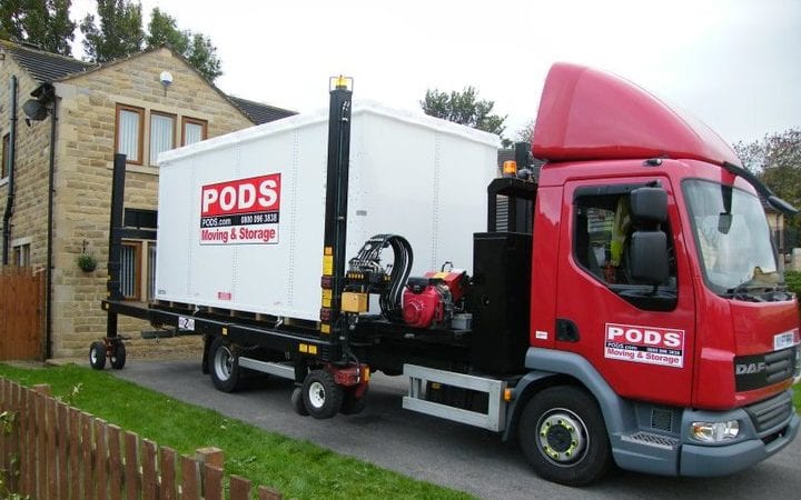 A PODS moving a storage truck is backed into a driveway and in the process of picking up a fully loaded portable storage container.