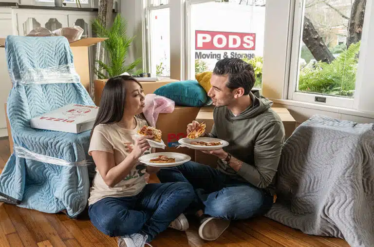 A man and woman are sitting on the floor of their house, eating pizza while they take a break from moving. The furniture around them is covered with moving blankets, and there are several packed moving boxes nearby. Through the window, a PODS moving container can be seen sitting in the driveway.