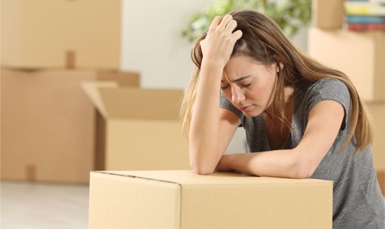 A young woman is leaning on a moving box with her head in her hand and a stressed expression on her face. There are several more moving boxes in the background.