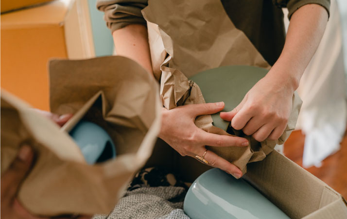 A close-up view of two people wrapping fragile dishes in brown paper before packing them away in cardboard moving boxes.