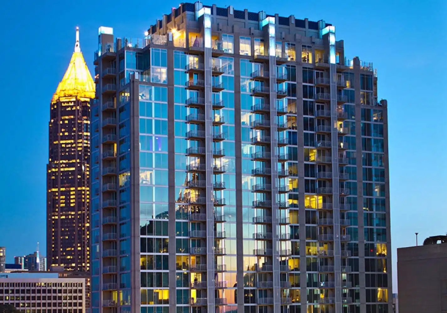 View of the top half of a luxury Atlanta apartment building at dusk. The sky is a dark blue, and the yellow lights of several units in the building are shining through the windows to the outside.