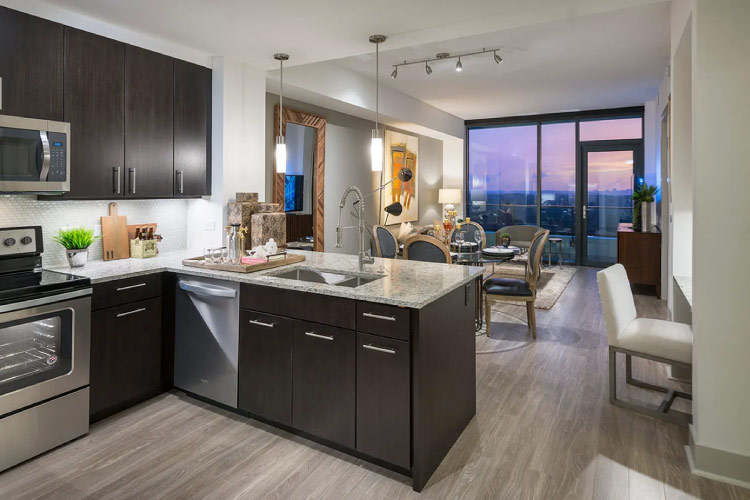 Interior view of an apartment at Hanover Buckhead Village. The kitchen features quartz countertops, and the adjacent living room has floor-to-ceiling windows with a sliding glass door that leads to a private balcony. The view outside is of a soft pink sunset over Buckhead and Midtown.