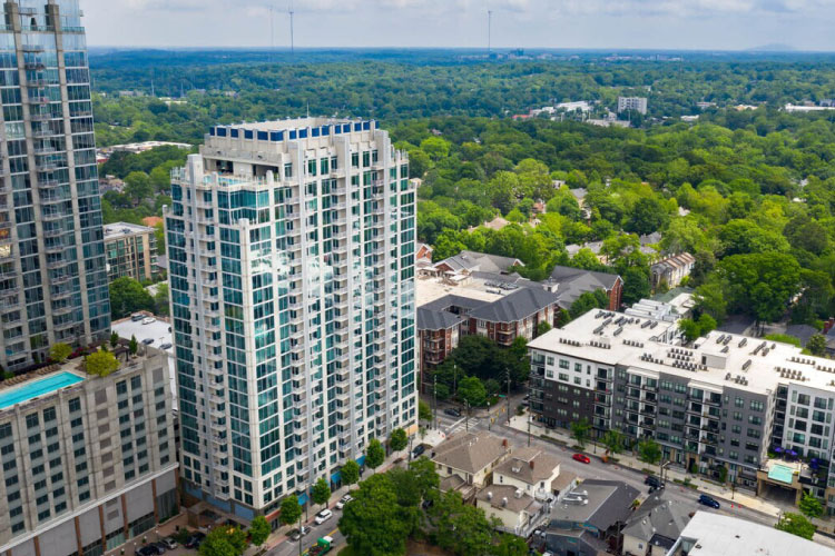 Aerial view of the luxury apartment building SkyHouse Buckhead in Atlanta, Georgia. There’s another tall building to the left, some shorter buildings to the right, and a lush green wooded area beyond them.