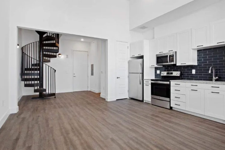Interior view of an empty apartment at Smith & Porter Apartments in Atlanta, Georgia. The apartment features white walls and wood flooring and an open kitchen with white cabinets and a black subway tile backsplash. In the entryway, beside the kitchen, is a black spiral staircase leading to the second level.