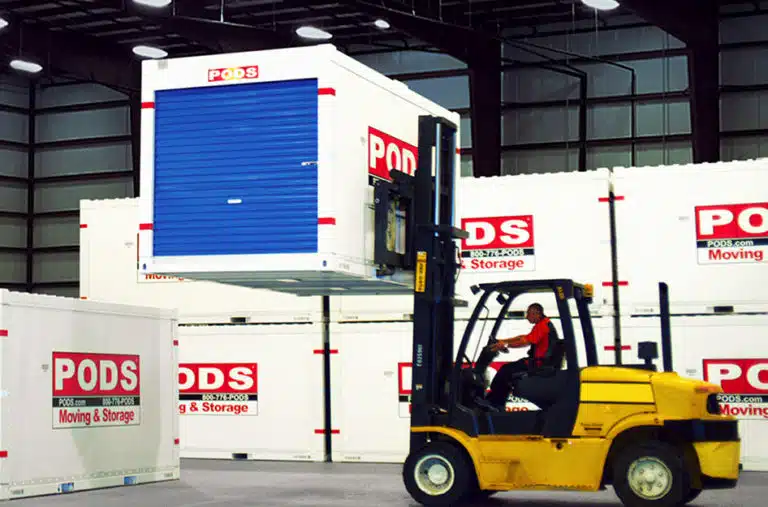 A forklift driver is lifting a PODS portable storage container to stack it above another container. In the background, several other PODS containers are stacked neatly against a PODS storage center wall.