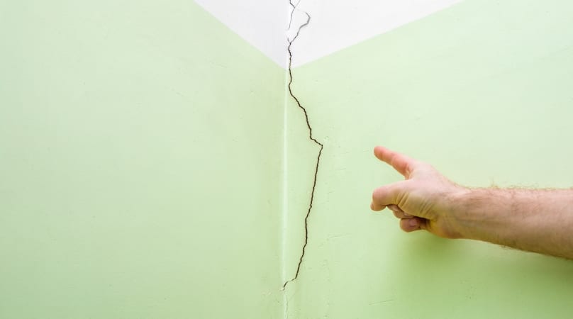 A crack runs down the middle of a green wall while a human finger points to the damage. 