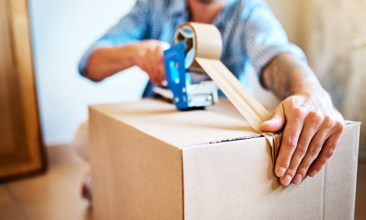 A man in a blue, button-up shirt is sitting on the floor, taping up a moving box that he just finished packing.