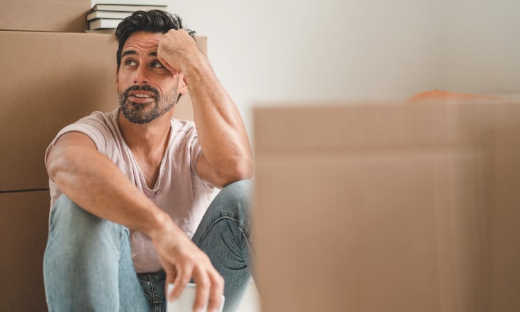 A man with a short, scruffy beard is sitting on the floor, leaning against a stack of moving boxes. He’s taking a coffee break on moving day.