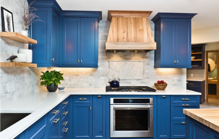 A modern farmhouse kitchen with navy blue shaker-style cabinets. The main counter is white and the island has a butcher block top. The vent over the stove is covered in wood to match the island. 
