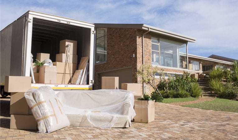 The back of a moving truck rental is open and the truck is partially loaded with moving boxes and other random things. Behind the truck, there are more boxes and furniture wrapped in plastic.