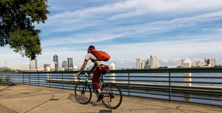 A cyclist is traveling along the waterfront, with the downtown Jacksonville skyline in the distance.