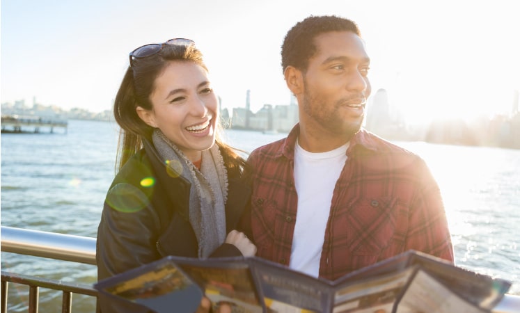 A happy couple laughs together as they explore their new city. They are standing by the water with the city skyline behind them. The man is holding a map and looking out.