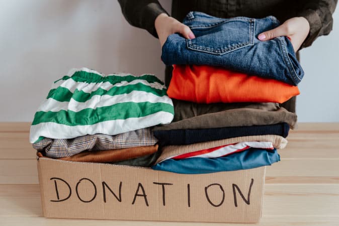 A woman is placing a stack of folded clothes into a shallow cardboard box labeled “DONATION.” There’s a variety of items in the box, including swim trunks, denim jeans, and plaid slacks.