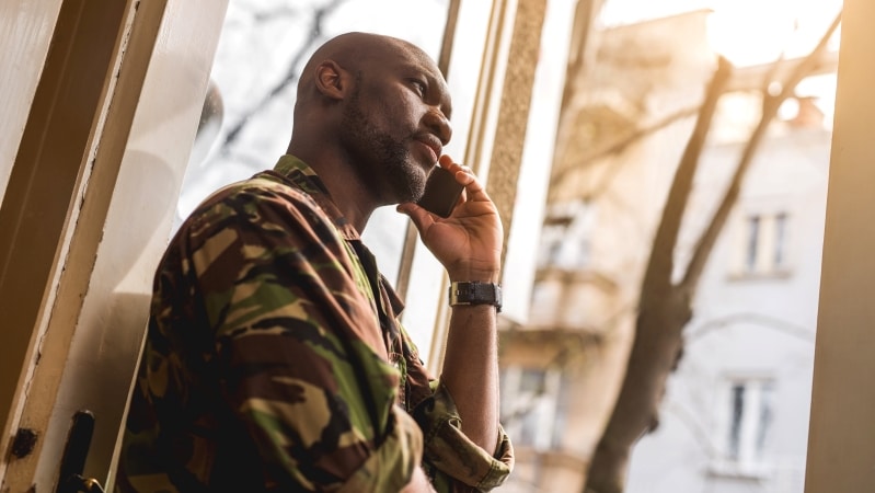 A military serviceman is standing in a doorway and speaking on his cell phone.