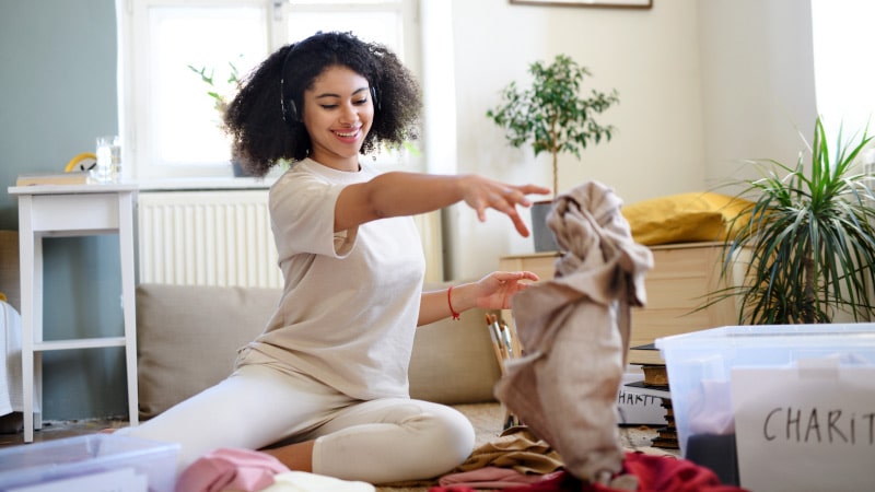 A motivated woman decluttering her belongings before moving. She is sitting on the floor and throwing a garment into a box labeled “CHARITY.”