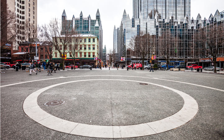 The center of Market Square in downtown Pittsburgh, PA. The bare concrete circle is surrounded by people, cars, and buildings.