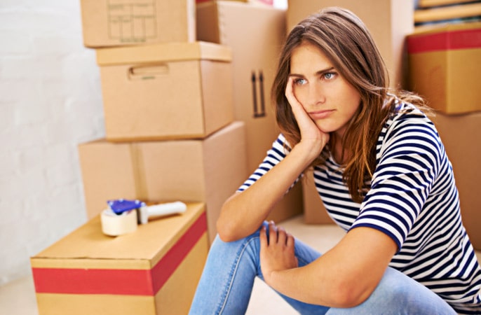 A woman with a tired expression on her face is sitting next to large stacks of packed moving boxes. She has her elbows propped up on her knees, and her chin resting on her hand.