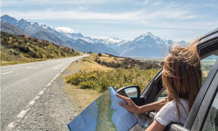 A woman pulled over on the side of the road, holding a map and looking at the road and mountains ahead.